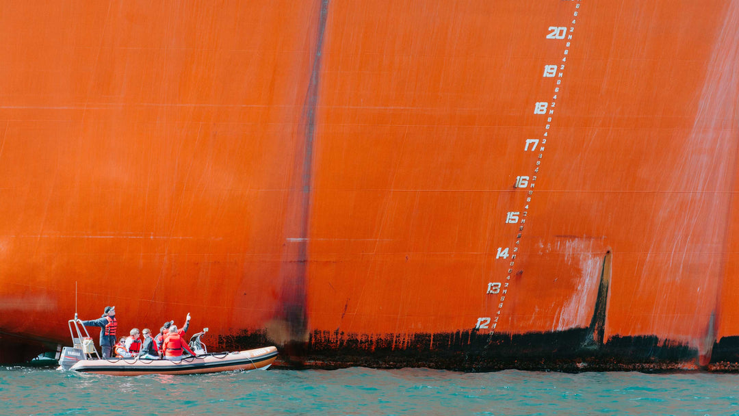 A large orange cargo ship sits behind a small inflatable boat illustrating the size difference between ships and boats